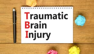 "TBI: Traumatic Brain Injury" written on a white note on a beautiful wooden table. A pen is also placed on the wooden background.