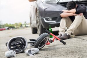 A desperate, scared, and stressed drunk driver sits with a bottle of beer in front of a crashed car, which has collided with a child's bike on the city road after an accident. 