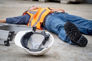 A construction site accident and injury emphasize the "Safety First" concept. A male worker lies unconscious on the ground, still wearing his safety helmet, after falling from a ladder.