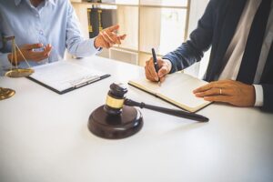 A judge's gavel is positioned prominently in the foreground, with a team of justice lawyers engaged in a meeting at a law firm in the background. 