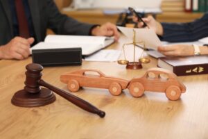 Closeup of wooden toy cars next to a judge's gavel on a lawyer's desk during a meeting in an office setting.