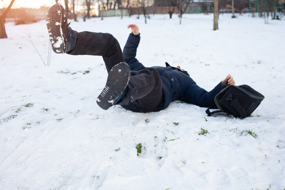 A man falls in the snow after slipping on ice, showing signs of injury. The winter scene highlights the risk of fractures, bruises, and dislocations due to slippery conditions.