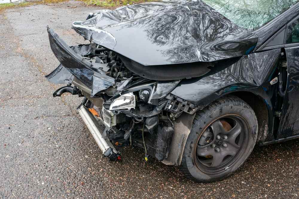 Black car with a crushed hood and torn-away front bumper sitting on wet pavement after a collision