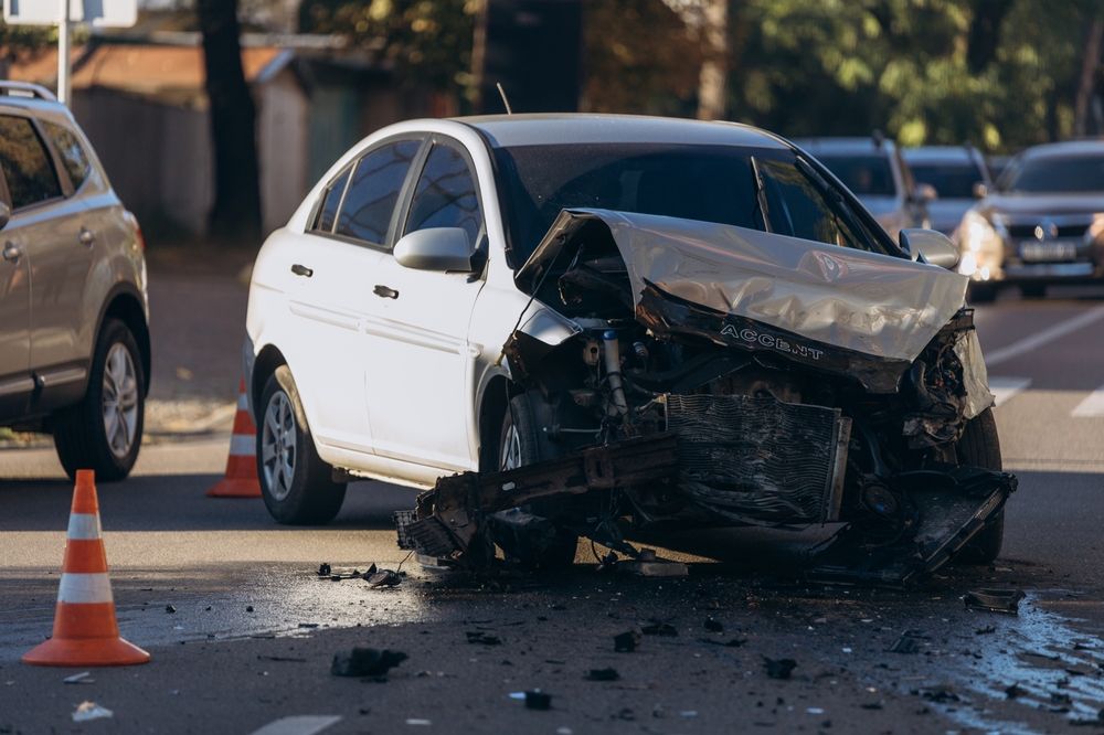 White sedan with a crushed front end surrounded by debris and traffic cones on a city street after a crash